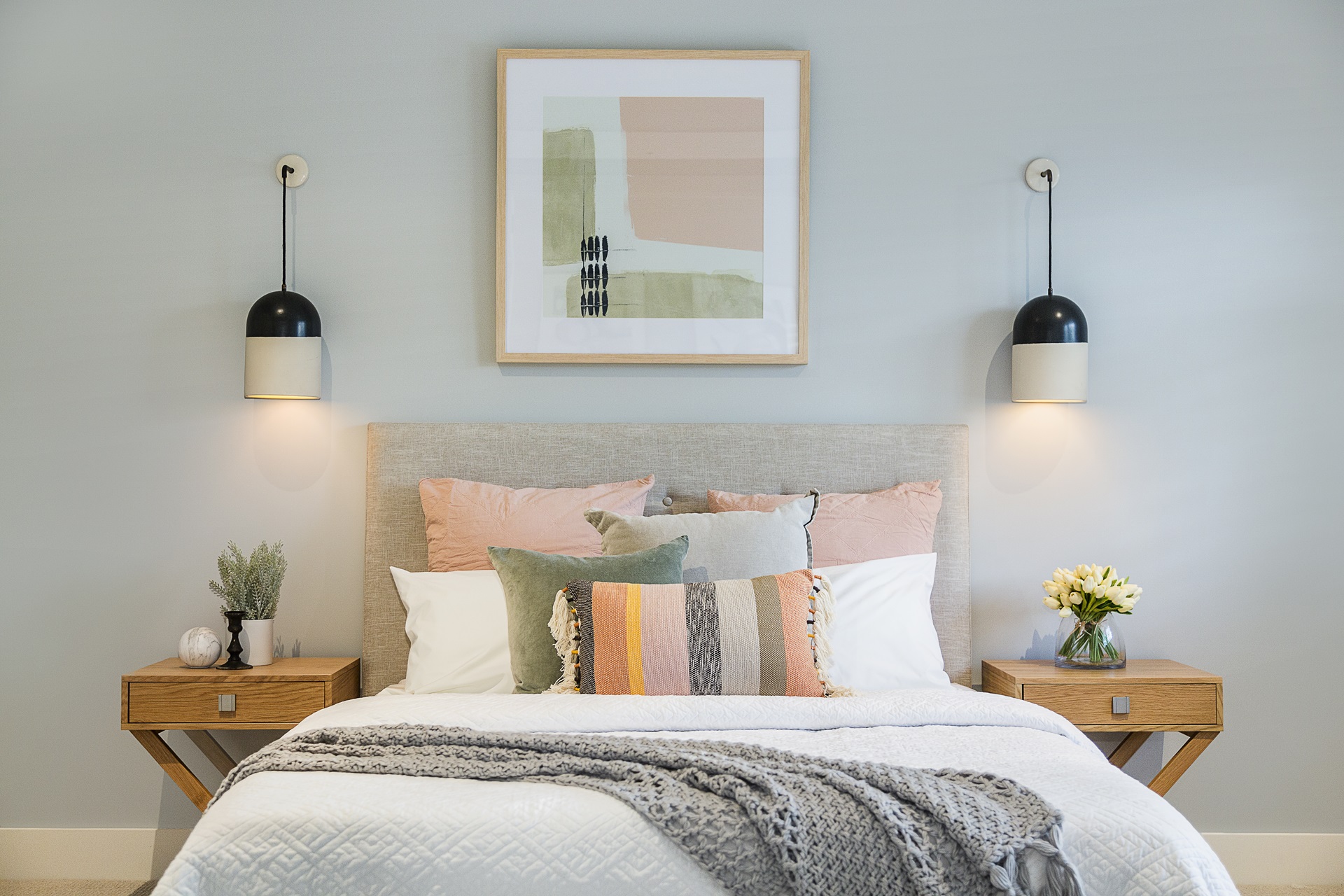 A neatly arranged bedroom with a bed featuring pastel and neutral pillows. Two wood nightstands hold lamps and decorations. A framed abstract artwork hangs above the bed.