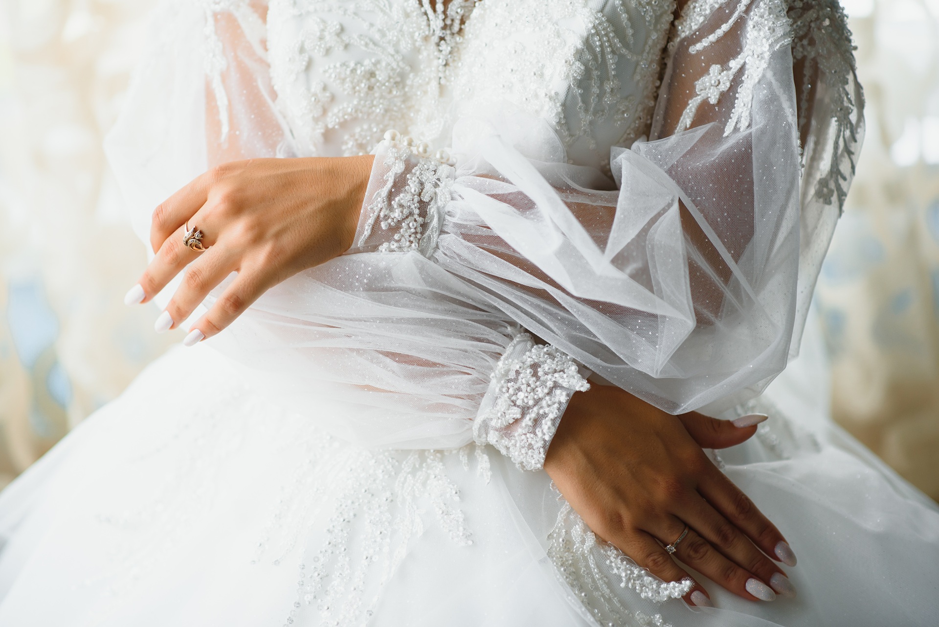 Close-up of a person in a white wedding dress with sheer, embellished sleeves, showing their hands with a ring on one finger.