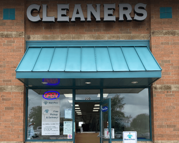 Storefront of a dry cleaners with a sign reading "CLEANERS." The door is open, displaying signs for free pickup and delivery. An "OPEN" sign is in the window.