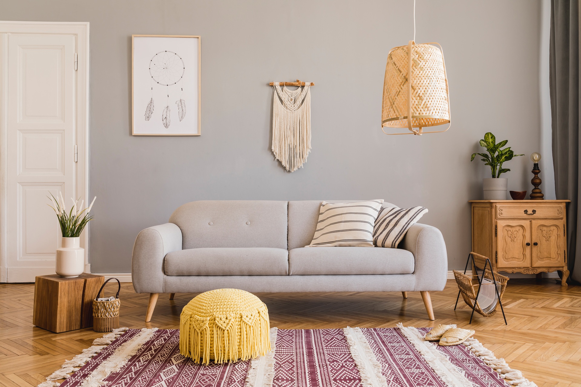 A modern living room with a light gray sofa, striped pillows, a wicker pendant lamp, wooden furniture, and wall art, featuring a patterned rug and a yellow pouf on a wooden floor.