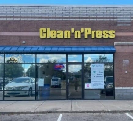 Storefront of a dry cleaners with a sign reading "CLEANERS." The door is open, displaying signs for free pickup and delivery. An "OPEN" sign is in the window.