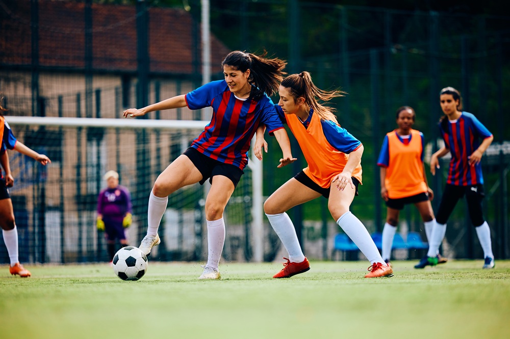 Two people are lying on massage tables, covered with dark towels. A person in a white robe stands nearby, holding a glass. The setting appears to be a spa or relaxation room.